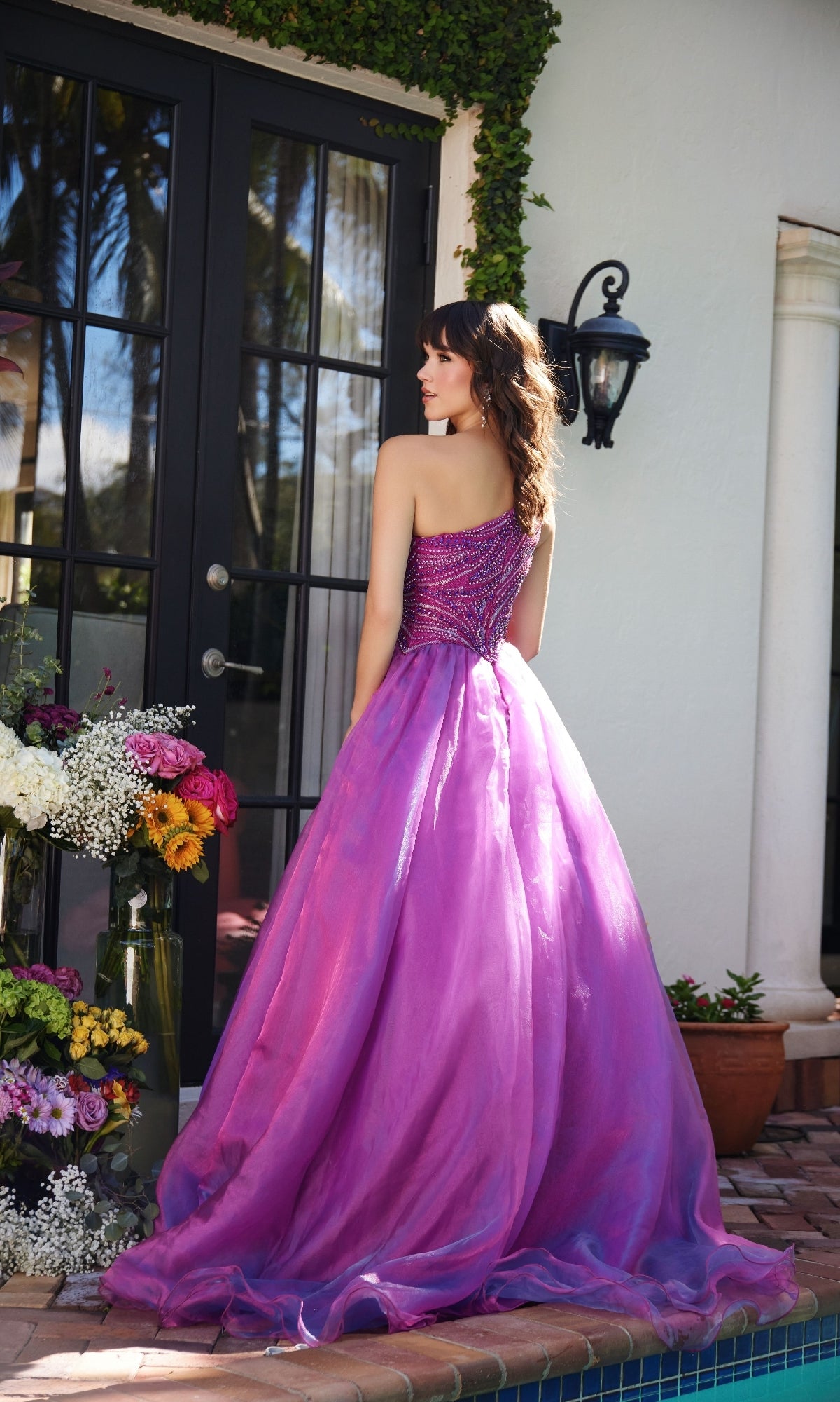 A woman in the Ava Presley 29258 one-shoulder prom ball gown stands by a glass door near a pool, surrounded by flower arrangements.